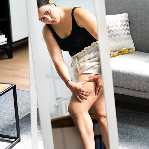 Frau schaut sich im Spiegel ihre Oberschenkel mit Cellulite an
