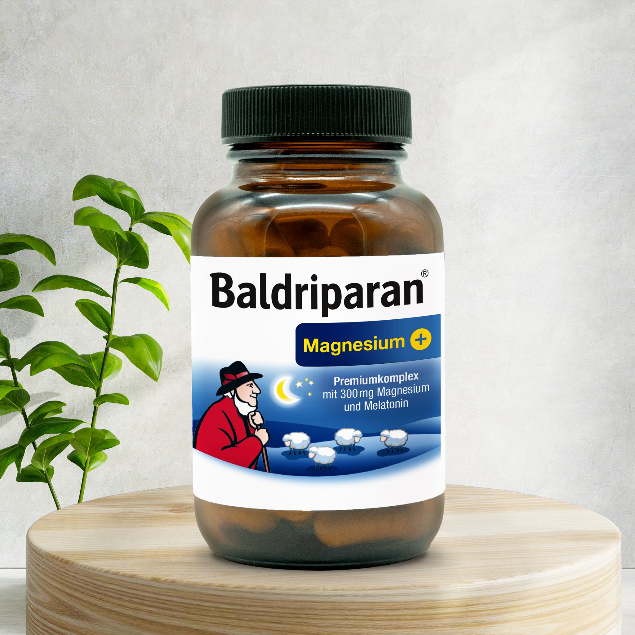 Braune Flasche Baldriparan® Magnesium+ mit Etikett: Premiumkomplex mit 300 mg Magnesium und Melatonin. Im Design: Hirte mit Schafen und Mondsichel. Die Flasche steht auf einem Holzpodest, daneben eine grüne Pflanze vor hellem Hintergrund.